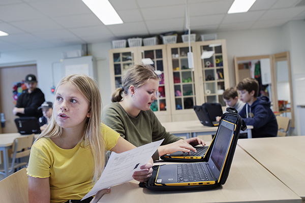 Två elever sitter vid bärbara datorer i ett klassrum. En av dem har ett papper i handen. I bakgrunden finns fler elever vid datorer.