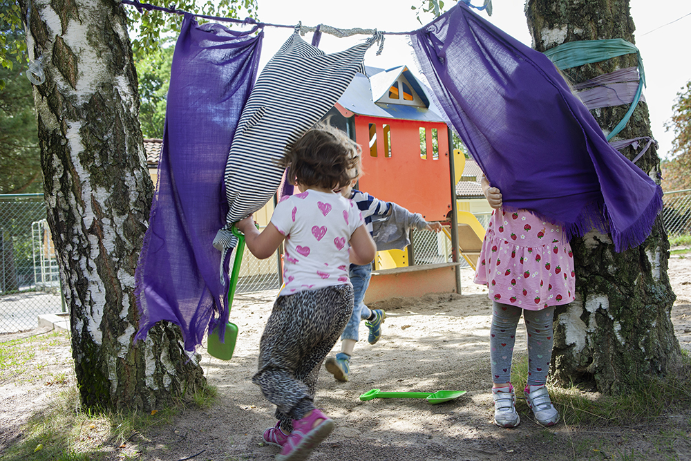 Förskolebarn leker under en tvättlina där färgglada tyger hänger.